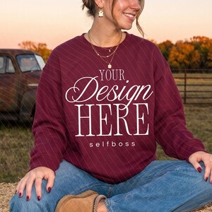 Op de afbeelding: Een bordeauxrood sweatshirt met lange mouwen en de witte tekst "YOUR Design HERE" en "selfboss" op de voorzijde. Het kledingstuk wordt gecombineerd met een blauwe spijkerbroek en beige suède instappers voor een casual uitstraling.