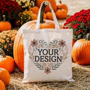 以下が含まれることがあります： 白いトートバッグに「YOUR DESIGN」の文字が花のハートの中にあります。バッグはカボチャ、菊、干し草のベールに囲まれ、秋の収穫をテーマにしています。