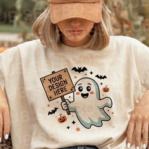 以下が含まれることがあります： 「YOUR DESIGN HERE」と書かれた看板を持つハロウィーンの幽霊のグラフィックが描かれたベージュのTシャツ。モデルは「PUMPKIN SPICE」が刺繍された茶色のコーデュロイベースボールキャップを着用しています。