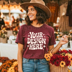 Può includere: Una maglietta bordeaux con la scritta bianca "YOUR DESIGN HERE", abbinata a jeans blu e un cappello marrone. Si tiene un cestino di vimini pieno di fiori rossi e gialli. Lo sfondo mostra un mercato dei fiori.