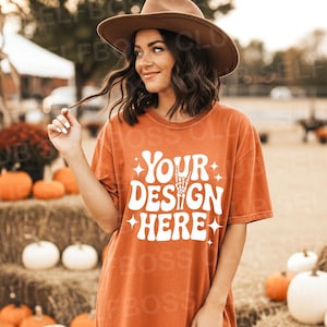 Pode incluir: Uma camiseta laranja com o texto branco "YOUR DESIGN HERE" e um gráfico de mão de esqueleto. A camisa é usada com leggings marrons e um chapéu marrom. Abóboras e fardos de feno ao fundo.