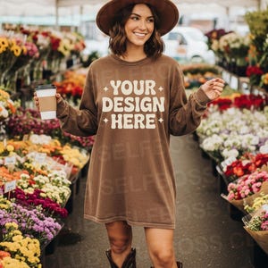 May include: A brown long-sleeve shirt dress with the text "YOUR DESIGN HERE" in white. The person in the image is wearing a brown hat and cowboy boots, holding a coffee cup, and standing in a flower market.