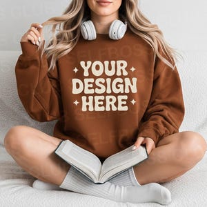 Puede incluir: Una sudadera marrón con el texto "YOUR DESIGN HERE" en una fuente retro. La persona lleva calcetines blancos y tiene auriculares blancos alrededor del cuello. Está sosteniendo un libro.