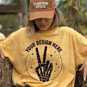 Pode incluir: Camiseta amarelo mostarda com um gráfico de mão de esqueleto fazendo o sinal de paz e o texto "YOUR DESIGN HERE". A modelo usa um boné de beisebol de veludo cotelê marrom com "PUMPKIN SPICE".