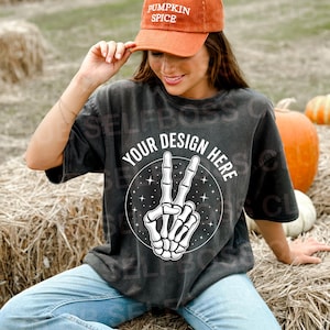Puede incluir: Camiseta gris oscuro con un gráfico de signo de la paz de la mano de un esqueleto y el texto "YOUR DESIGN HERE". La persona lleva una gorra de béisbol de pana naranja quemado con el texto "PUMPKIN SPICE". Calabazas y balas de heno en el fondo.