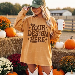 Puede incluir: Camiseta de manga larga color mostaza con el texto "YOUR DESIGN HERE" en marrón. La camiseta se usa con una gorra verde y botas blancas. Calabazas y balas de heno en el fondo, creando un tema otoñal.