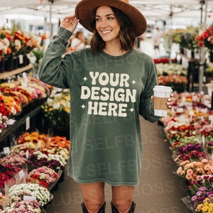Peut inclure: Robe-t-shirt vert olive à manches longues avec le texte "YOUR DESIGN HERE" en blanc. La personne porte un chapeau marron et des bottes noires, tenant une tasse de café. Le fond montre un marché aux fleurs avec diverses fleurs colorées.