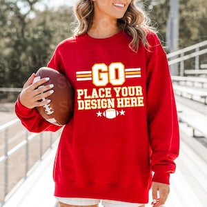 May include: A red sweatshirt with the text "GO PLACE YOUR DESIGN HERE" in a yellow and white design. The sweatshirt is being held by a person holding a brown football.