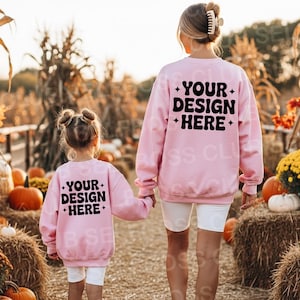May include: Two pink sweatshirts with the text "YOUR DESIGN HERE" on the back. A woman and a child are walking hand-in-hand through a pumpkin patch. The sweatshirts are long-sleeved.