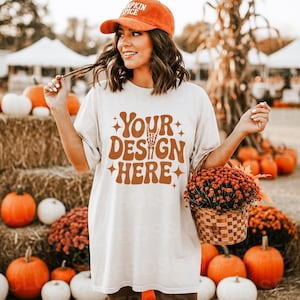 May include: A cream-colored t-shirt with the text "YOUR DESIGN HERE" in brown, worn with an orange cap that says "PUMPKIN SPICE". The scene includes pumpkins, hay bales, and a basket of flowers, evoking an autumn theme.