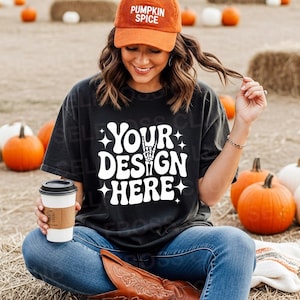 May include: A woman wearing a black t-shirt with the text "YOUR DESIGN HERE" in white, a corduroy orange cap with "PUMPKIN SPICE" text, and blue jeans. She is holding a coffee cup and sitting on a hay bale with pumpkins in the background.