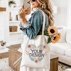May include: A cream-colored tote bag with a floral heart design and the text "YOUR DESIGN". The bag is filled with sunflowers. The person is wearing sunglasses and a denim jacket.