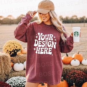 May include: A maroon long-sleeve sweatshirt with the text "YOUR DESIGN HERE" in white. The image features pumpkins, hay bales, and a person wearing a baseball cap and holding a coffee cup.