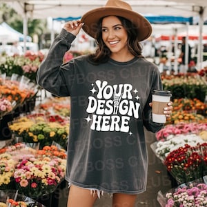 May include: Dark gray long-sleeve shirt with the text "YOUR DESIGN HERE" in white. The shirt is worn with a brown hat and boots. The background features a flower market with various colorful blooms.