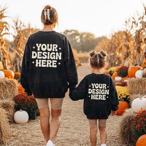 Può includere: Felpe nere con la scritta "YOUR DESIGN HERE" in bianco. Una madre e una bambina camminano in un campo di zucche. Le felpe sono un set coordinato, perfetto per l'autunno.