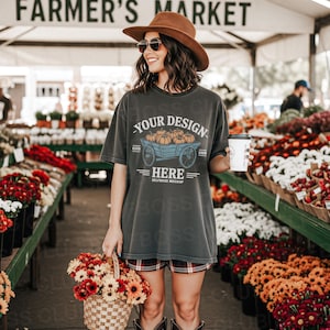 May include: A woman at a farmer's market wears a gray t-shirt with a pumpkin cart design and the text "YOUR DESIGN HERE". She holds a coffee cup and a basket of flowers. The market features flower displays and the sign "FARMER'S MARKET".