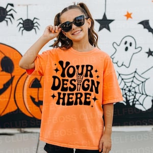 May include: An orange t-shirt with the text "YOUR DESIGN HERE" in black, featuring a skeleton hand. The shirt is worn by a person wearing sunglasses, with a Halloween-themed background of pumpkins, spiders, and ghosts.