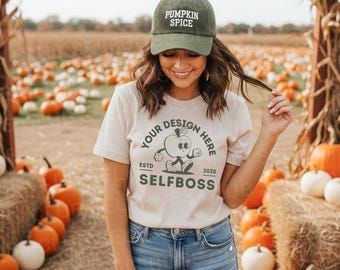 May include: A woman wearing a green "Pumpkin Spice" baseball cap and a cream-colored t-shirt with a graphic that says "Your Design Here" and "Selfboss". She is in a pumpkin patch, surrounded by pumpkins and hay bales.