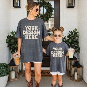 Könnte beinhalten: Zwei Personen tragen passende graue T-Shirts mit dem Text "YOUR DESIGN HERE" in weißer Retro-Schrift. Beide tragen Sonnenbrillen und Stiefel. Das Bild wurde vor einem weißen Backsteingebäude aufgenommen.