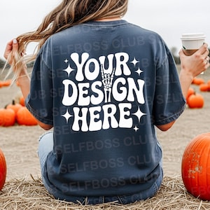 May include: Dark blue t-shirt with the white text "YOUR DESIGN HERE" and a skeleton hand graphic. The image shows the shirt being worn outdoors with pumpkins in the background.