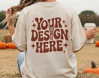 May include: A beige t-shirt with the text "YOUR DESIGN HERE" in a brown, retro font with a skeleton hand gesture. The shirt is worn outdoors with pumpkins and hay bales in the background.