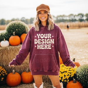 May include: A maroon hoodie with the text "YOUR DESIGN HERE" in white. The hoodie has a front pocket and a drawstring hood. The image is set outdoors with pumpkins and autumn foliage.
