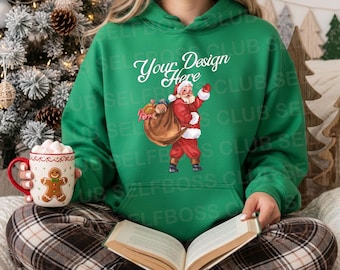 May include: A green hoodie with a Santa Claus graphic carrying a sack of toys. The hoodie has the text "Your Design Here" in white script. A person is holding a mug with a gingerbread man design and a book.