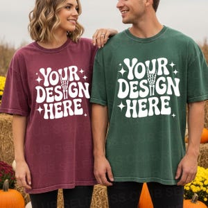 May include: Two oversized t-shirts, one burgundy and one dark green, each with the text "YOUR DESIGN HERE" in white, featuring a skeleton hand gesture. The shirts are worn by two people standing in front of a fall harvest scene.