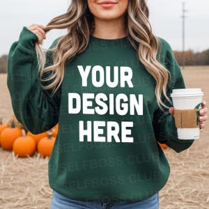May include: A dark green sweatshirt with the text "YOUR DESIGN HERE" in white. The sweatshirt is being held by a person. A coffee cup with a brown sleeve is in the person's hand. Pumpkins are in the background.