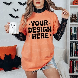 May include: Orange t-shirt with the text "YOUR DESIGN HERE" in black. The model is holding a white ghost-shaped mug. The background includes Halloween decorations like bats and a pumpkin.