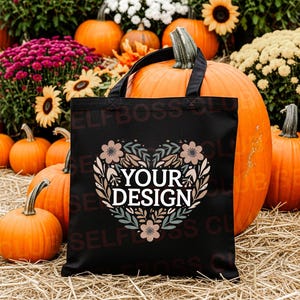 May include: A black tote bag with a floral heart design and the text "YOUR DESIGN" is displayed in front of pumpkins, flowers, and hay. The pumpkins are various sizes and shapes, with orange skin and green stems.