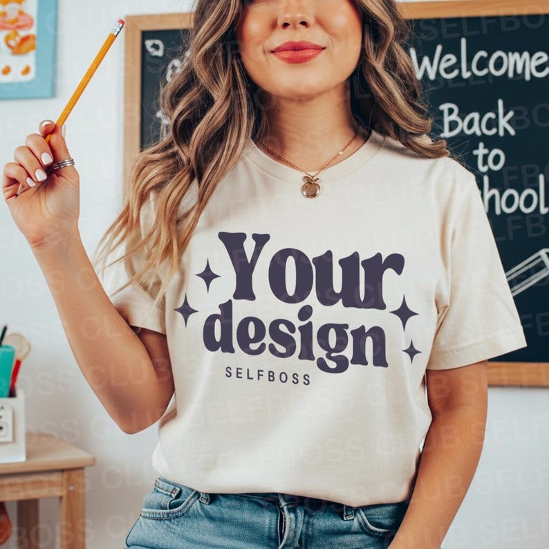 May include: Cream-colored t-shirt with the text "Your design" in a dark blue, stylized font, with the word "SELFBOSS" below. The shirt is short-sleeved and worn by a person. A pencil is held in the person's hand.
