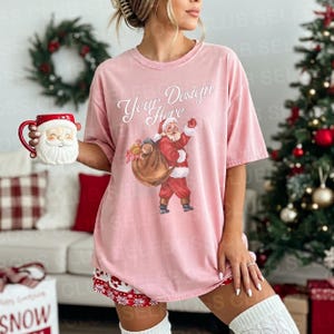 May include: A light pink t-shirt with a Santa Claus graphic and the text "Your Design Here." The Santa illustration features a red suit and a bag of toys. A Santa mug is held in the left hand. The theme is Christmas.