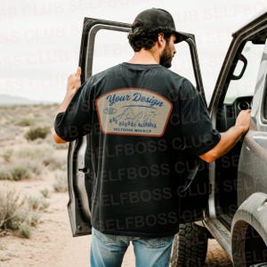 May include: Black t-shirt with a patch on the back that says "Your Design" above a mountain and tree graphic. The shirt is worn by a person standing next to a vehicle in a desert landscape. The patch also says "SELFBOSS MOCKUP".