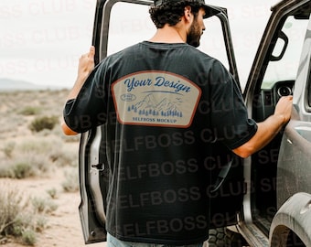 May include: Black t-shirt with a patch on the back featuring the text "Your Design" above a mountain and tree graphic. The shirt is worn by a person standing next to a vehicle in a desert landscape. The patch also says "SELFBOSS MOCKUP".