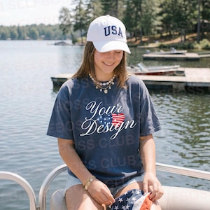 Pode incluir: Uma mulher usando um boné de beisebol branco com "USA" e uma camiseta azul marinho com "Your Design" e uma bandeira americana. Ela está sentada em um barco perto de um lago.