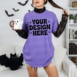 May include: A lavender t-shirt with the text "YOUR DESIGN HERE" in black. The model is wearing a black lace sleeve shirt underneath. A white ghost-shaped mug is held in the left hand. Halloween decorations, including bats, are in the background.