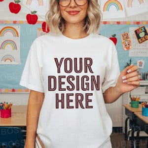 May include: A white t-shirt with the text "YOUR DESIGN HERE" in a maroon color. The shirt is short-sleeved and appears to be made of a soft material. The background includes classroom decorations with rainbows and apples.