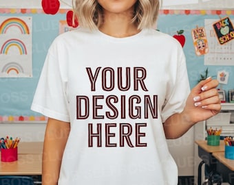 May include: A white t-shirt with the text "YOUR DESIGN HERE" in a maroon color. The shirt is short-sleeved and appears to be made of a soft material. The background includes classroom decorations with rainbows and apples.