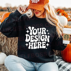 Könnte beinhalten: Schwarzes Sweatshirt mit dem weißen Text "YOUR DESIGN HERE", mit einer Handgeste und Sternen. Die Person trägt eine orangefarbene Cord-Mütze mit der Aufschrift "PUMPKIN SPICE". Der Hintergrund umfasst Kürbisse und Heu.