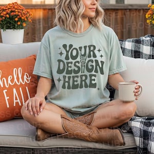 May include: A light green t-shirt with the text "YOUR DESIGN HERE" in dark green, featuring a hand skeleton graphic. The person is wearing brown cowboy boots and holding a beige mug. An orange cushion with "hello FALL" is visible.