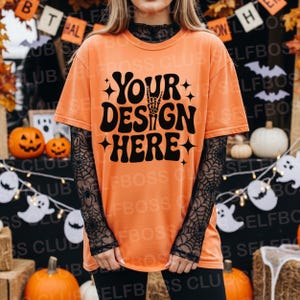 May include: Orange t-shirt with the text "YOUR DESIGN HERE" in a retro font, featuring a skeleton hand. The shirt is layered over a black lace top. Halloween-themed apparel.