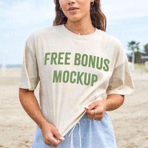 Puede incluir: Camiseta color crema con el texto "FREE BONUS MOCKUP" en verde. La camiseta est&aacute; siendo sostenida por una persona. La persona lleva pantalones cortos azules y est&aacute; en una playa.