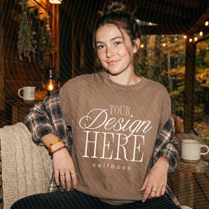 May include: A brown crewneck sweatshirt featuring the white text "YOUR Design HERE" and "selfboss" printed on the front. The garment is worn layered over a plaid button-down shirt, showcasing a casual style with visible fabric texture.