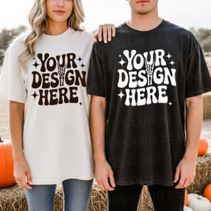 May include: Two short-sleeved t-shirts, one cream and one black, each with the text "YOUR DESIGN HERE" in a retro font. The design includes a hand skeleton making a rock-and-roll gesture. The shirts are worn by two people.