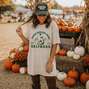 Puede incluir: Una mujer lleva una camiseta beige con el texto "YOUR DESIGN HERE" y un dibujo de una manzana de dibujos animados. También lleva una gorra de béisbol de pana verde que dice "PUMPKIN SPICE". Hay calabazas y balas de heno en el fondo.