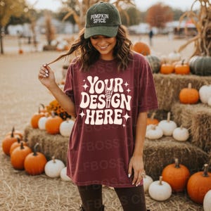 May include: A maroon t-shirt with the text "YOUR DESIGN HERE" in white, paired with a green baseball cap that says "PUMPKIN SPICE". The image is set outdoors with pumpkins and hay bales in the background.