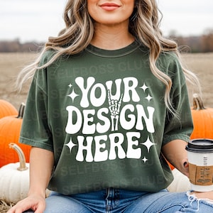 May include: Dark green t-shirt with the text "YOUR DESIGN HERE" in white, featuring a skeleton hand gesture. The shirt is worn with blue jeans. A coffee cup with a brown sleeve is held in the hand.