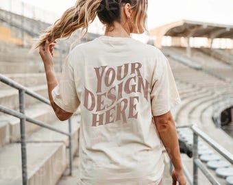 May include: A woman wearing a cream-coloured t-shirt with the text "YOUR DESIGN HERE" in brown lettering on the back. She is wearing brown shorts and has her hair pulled back in a ponytail.