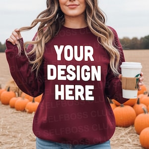 Op de afbeelding: Bordeaux sweatshirt met de witte tekst "YOUR DESIGN HERE". De persoon houdt een koffiekopje vast met een bruine mouw. Pompoenen zijn zichtbaar op de achtergrond.